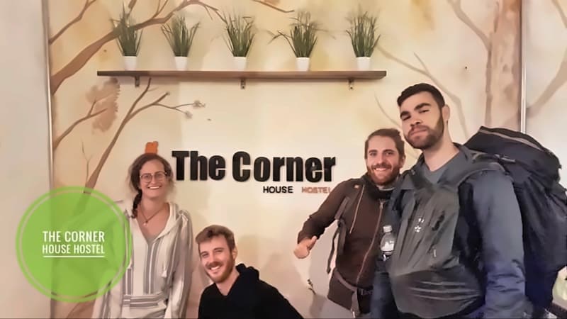 Four backpackers, two still carrying big packs, grinning beside the “The Corner” logo under a shelf of potted grasses