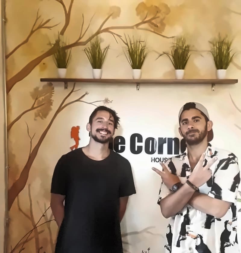 Two travellers posing in front of the painted branches of the reception mural, one crossing his arms in a double peace sign
