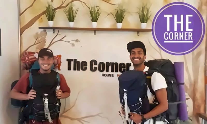 Two backpackers in caps, front and back packs on, smiling at reception in front of the logo wall