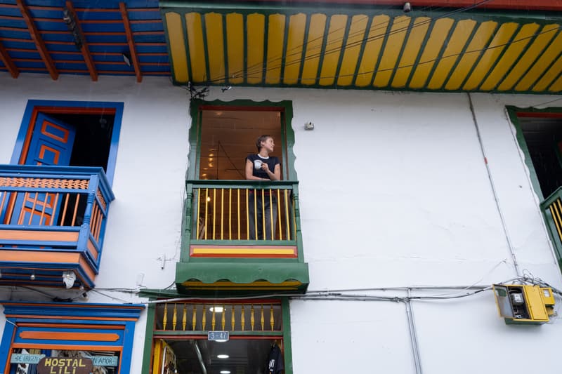 A guest with a coffee cup leaning on a green-and-yellow wooden balcony on the white façade, above blue painted doors