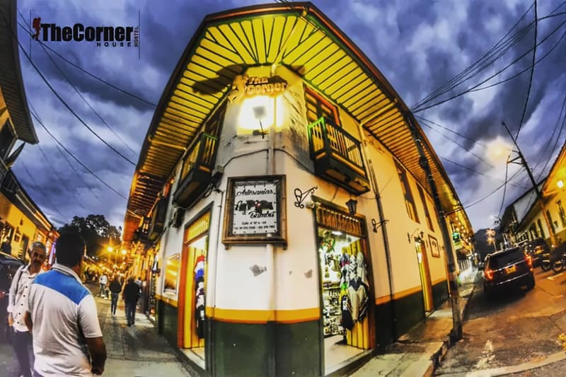 The Corner House at dusk: a white two-storey corner building with yellow painted eaves, green-and-yellow trim and wooden balconies, a lit sign at the corner and people walking on Salento’s main street