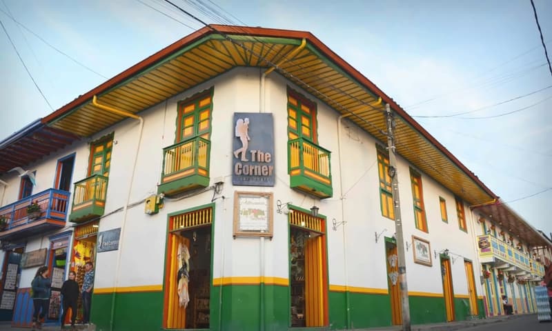 The corner building in daylight: white walls, a yellow eave with red trim, green-and-yellow balconies, a vertical “The Corner” sign and a green base with a yellow stripe