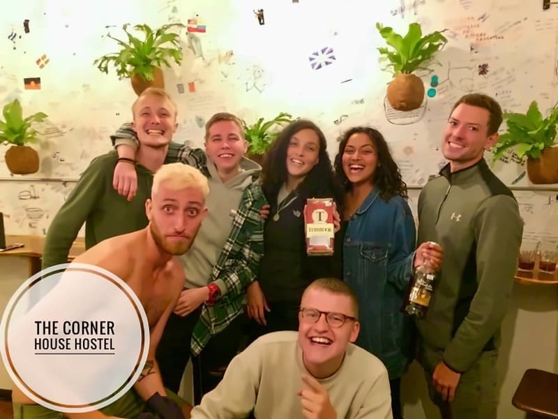 Seven smiling travellers posing together in front of the message wall, hanging plants and painted flags around them