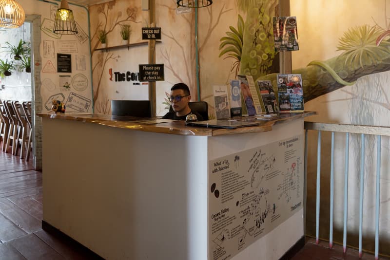 The reception desk today: the botanical mural, the “The Corner” logo, a brochure rack and a staff member at the counter