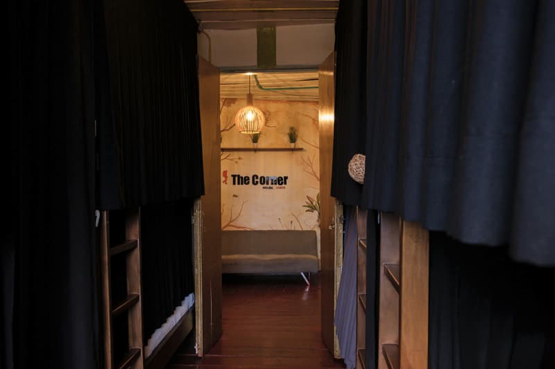 Large mixed dorm: view between curtained bunks towards the lounge, where the “The Corner” logo wall is visible
