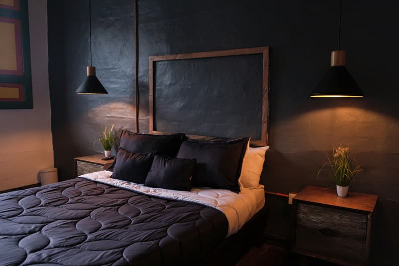 Deluxe Double Room: double bed with dark quilt and cushions, two black pendant lamps and small bedside tables against a dark wall