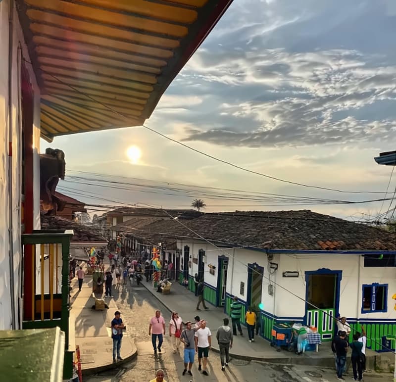 View from a first-floor balcony down Salento’s main street at sunset: white houses with blue and green doors, a palm on the skyline and people walking below