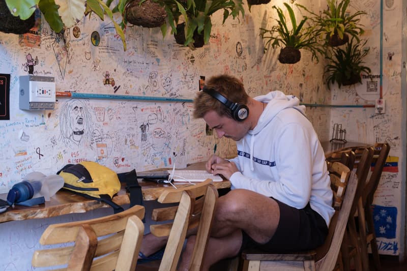 A guest with headphones writing at a wooden table beside the message wall, kokedama plants hanging above